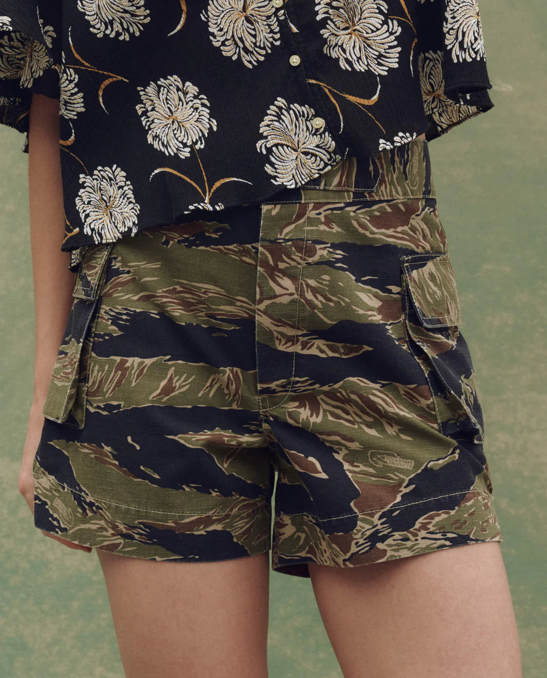 A person wears The Cargo Fatigue Short by The Great Inc. in green and brown camo with cargo pockets, paired with a black shirt featuring a white floral pattern. The shot highlights the shorts and torso, with a blurred background.