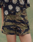 A person wears The Cargo Fatigue Short by The Great Inc. in green and brown camo with cargo pockets, paired with a black shirt featuring a white floral pattern. The shot highlights the shorts and torso, with a blurred background.