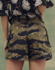 A person wears The Great Inc. The Cargo Fatigue Short in green and brown cotton camo with cargo pockets, paired with a black top featuring a white floral pattern, standing against a muted green background.