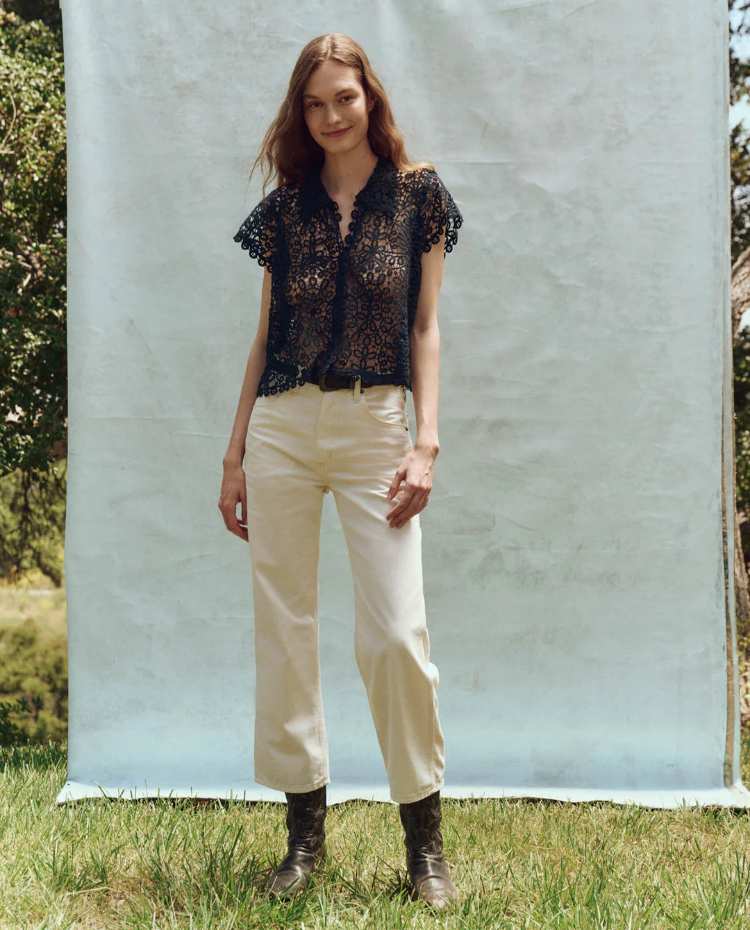 In front of a light blue backdrop, a woman stands outdoors on the grass wearing The Lattice Top by The Great Inc., featuring scalloped lace edges. She's paired it with cream pants and brown boots, her hands resting by her sides as she smiles softly, surrounded by trees and greenery.