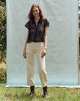 In front of a light blue backdrop, a woman stands outdoors on the grass wearing The Lattice Top by The Great Inc., featuring scalloped lace edges. She's paired it with cream pants and brown boots, her hands resting by her sides as she smiles softly, surrounded by trees and greenery.