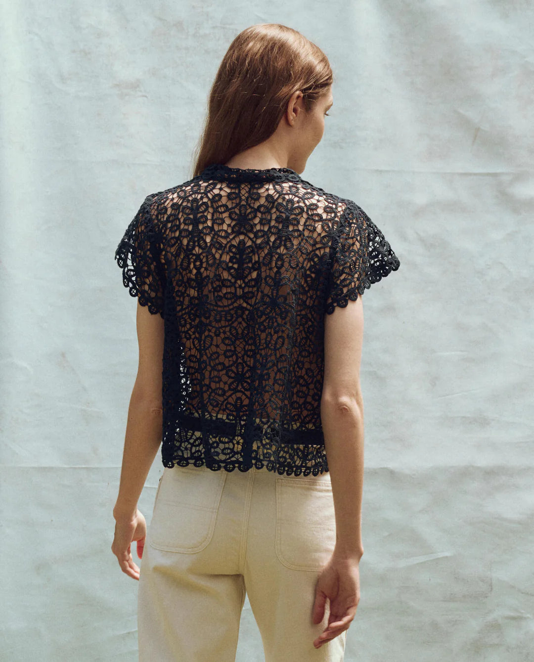 A person with long hair stands with their back to the camera, wearing The Lattice Top by The Great Inc., a black, intricately crocheted piece with scalloped lace edges, paired with light-colored pants against a plain, light background.