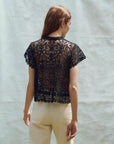 A person with long hair stands with their back to the camera, wearing The Lattice Top by The Great Inc., a black, intricately crocheted piece with scalloped lace edges, paired with light-colored pants against a plain, light background.