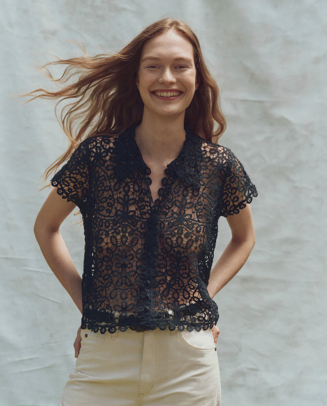 A smiling person with long hair poses against a textured gray backdrop wearing The Lattice Top by The Great Inc., featuring scalloped lace edges, paired with light-colored pants, and casually tucking their hands into their pockets.