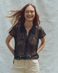 A smiling person with long hair poses against a textured gray backdrop wearing The Lattice Top by The Great Inc., featuring scalloped lace edges, paired with light-colored pants, and casually tucking their hands into their pockets.