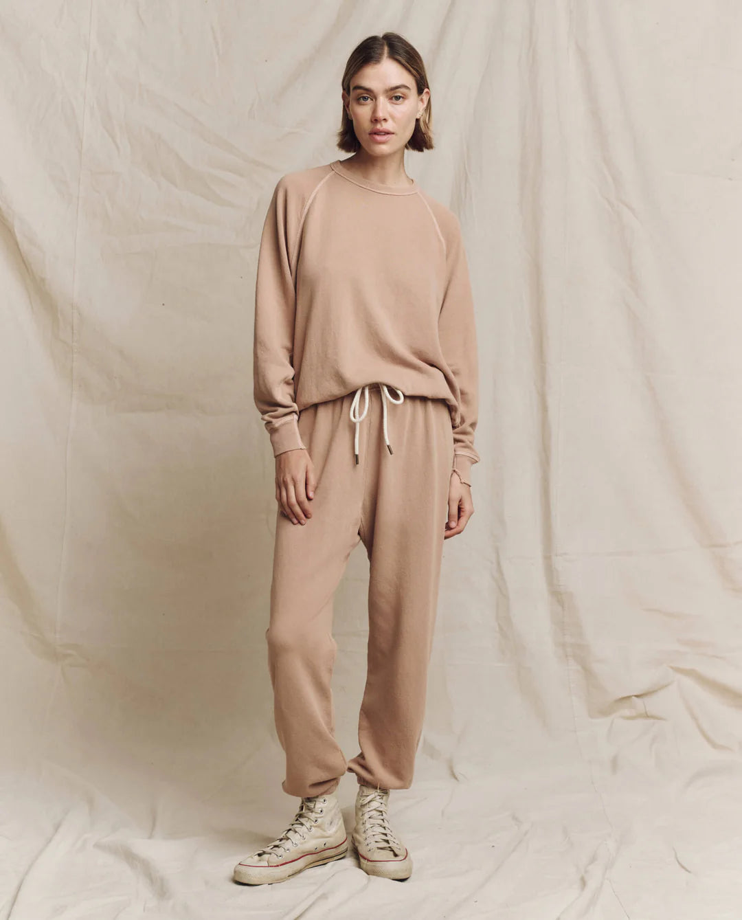 A person stands against a light beige fabric backdrop, dressed in coordinated beige athletic wear, including The Stadium Sweatpant by The Great Inc. Their short hair enhances the casual vibe, while white high-top sneakers complete the look. With hands relaxed at their sides, they perfectly capture the effortless style of Los Angeles.