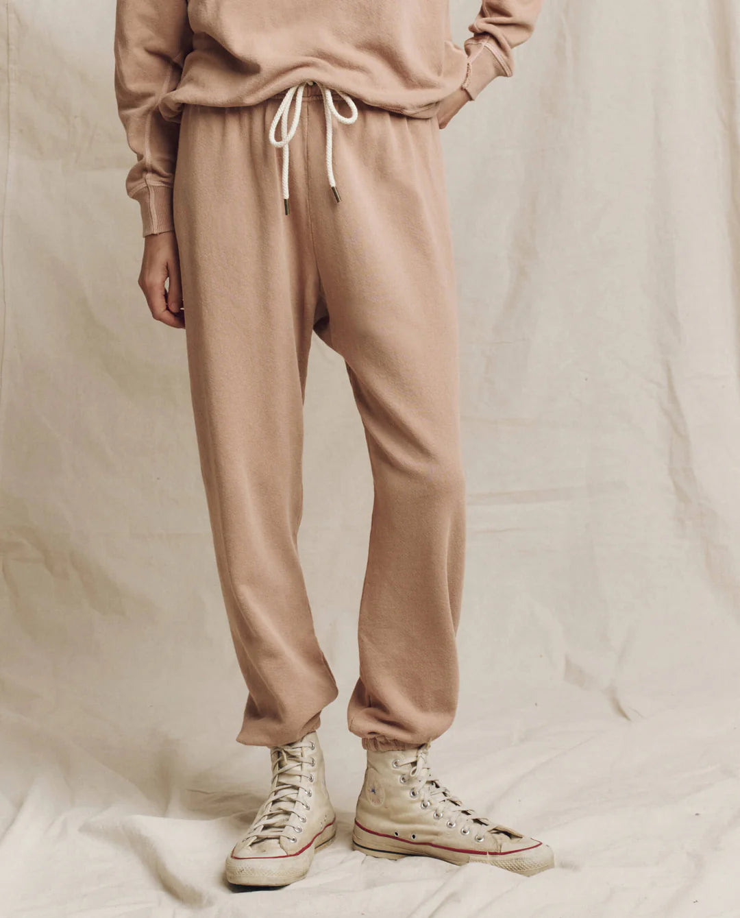 A person wearing The Great Inc.'s The Stadium Sweatpant in handcrafted beige, along with white high-top sneakers, stands against a neutral fabric backdrop. The outfit, reminiscent of vintage athletic wear, features drawstring pants and a 100% cotton sweatshirt.