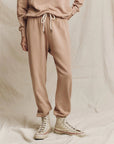 A person wearing The Great Inc.'s The Stadium Sweatpant in handcrafted beige, along with white high-top sneakers, stands against a neutral fabric backdrop. The outfit, reminiscent of vintage athletic wear, features drawstring pants and a 100% cotton sweatshirt.