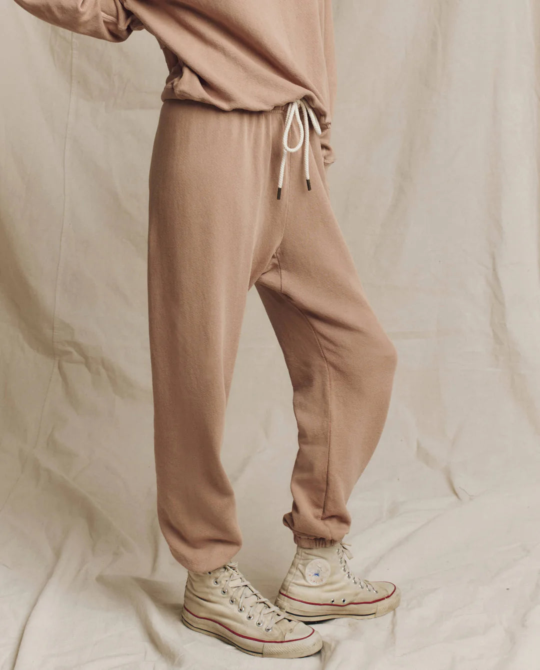 A person wearing The Stadium Sweatpant by The Great Inc. in beige and high-top sneakers poses against a cream backdrop. The joggers feature a drawstring waist and elastic cuffs, while the light-colored sneakers have a slightly worn appearance. Their relaxed stance exudes casual charm, evoking vintage athletic wear vibes.