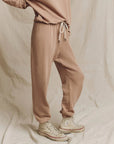 A person wearing The Stadium Sweatpant by The Great Inc. in beige and high-top sneakers poses against a cream backdrop. The joggers feature a drawstring waist and elastic cuffs, while the light-colored sneakers have a slightly worn appearance. Their relaxed stance exudes casual charm, evoking vintage athletic wear vibes.