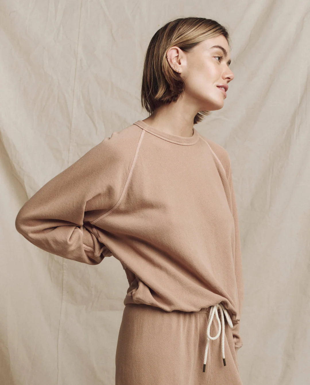 A person with short hair is wearing The College Sweatshirt by The Great Inc., in light brown, featuring a wild panther graphic, along with matching drawstring pants. They are standing sideways with one hand on their hip against a beige fabric backdrop.