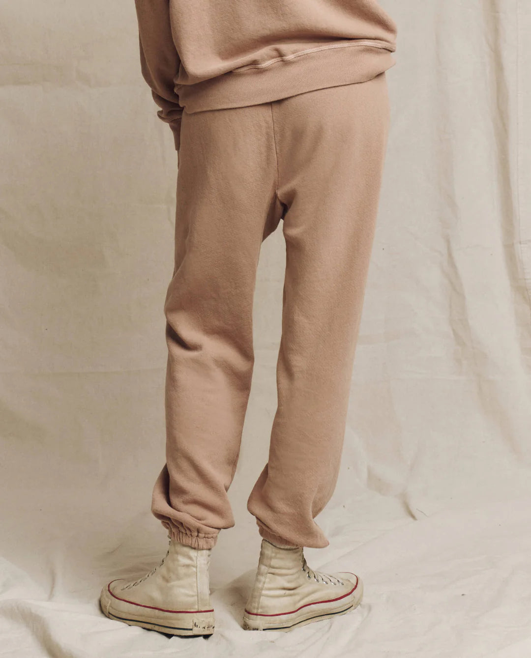 A person wearing The Stadium Sweatpant by The Great Inc. along with high-top off-white sneakers, reminiscent of vintage athletic wear, stands on a neutral fabric backdrop.