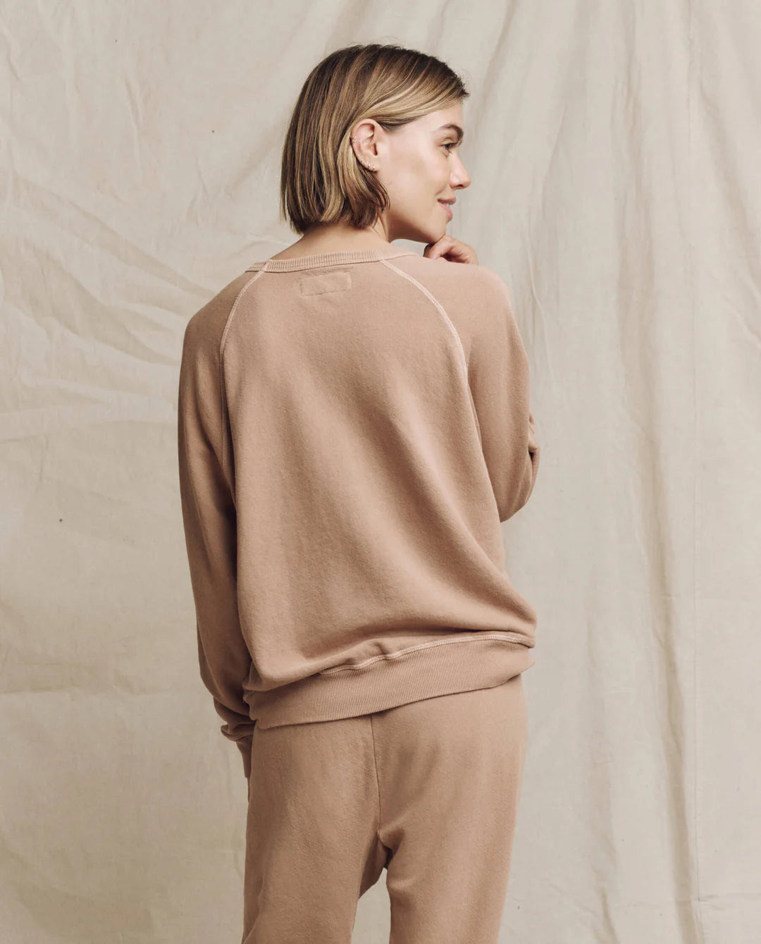 A person with short hair is standing in front of a beige backdrop, dressed in a light brown ensemble from The Great Inc. They are wearing The College Sweatshirt and matching pants featuring a Wild Panther Graphic. With a calm expression and sideways stance, they exude an effortless charm.