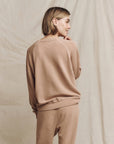 A person with short hair is standing in front of a beige backdrop, dressed in a light brown ensemble from The Great Inc. They are wearing The College Sweatshirt and matching pants featuring a Wild Panther Graphic. With a calm expression and sideways stance, they exude an effortless charm.