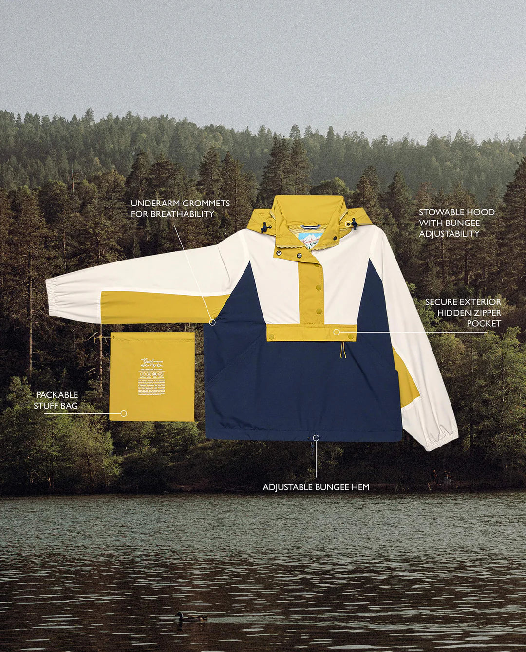 The Crestline Pullover by The Great Inc., set against a forest backdrop with a serene lake, is your adventure essential. This color-block windbreaker in yellow, white, and navy features recycled fabrics, underarm grommets, stowable hood, hidden zipper pocket, adjustable bungee hem, and packable stuff bag.