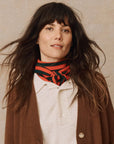 A person with long brown hair stands against a plain background, wearing The Cashmere Neckerchief by The Great Inc. in luxurious red and black stripes, paired with a white shirt and brown cardigan. The slightly tousled hair enhances their neutral expression.