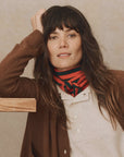 A person with long, dark hair and bangs leans against a wooden object, adorned in a brown cardigan from The Great Inc., paired with a beige shirt. They add a touch of coziness by wearing The Cashmere Neckerchief in red stripes around their neck. The textured neutral backdrop completes the scene.