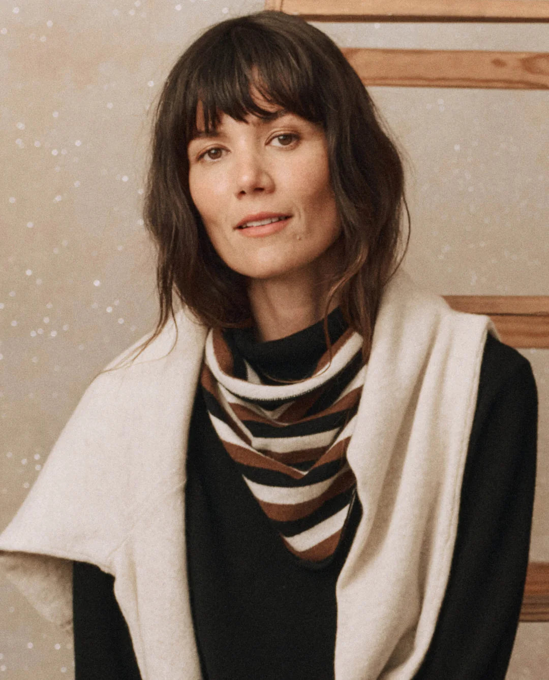 A person with long dark hair and bangs wears a dark top paired with The Cashmere Neckerchief from The Great Inc., and a sweater draped over their shoulders. They stand in front of a wooden structure, framed by a softly lit, speckled background.