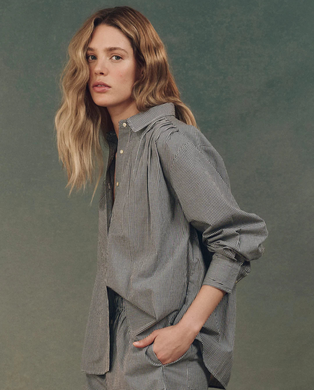 A person with long, wavy hair poses against a textured, greenish-blue backdrop wearing The Society Top by The Great Inc. They have a serious expression and their right hand is in their pocket.