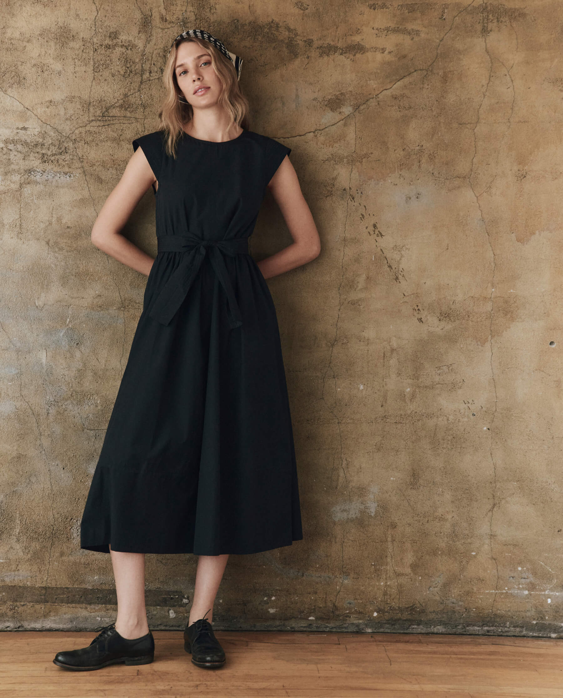 A woman with wavy hair and a relaxed expression stands against a textured beige wall wearing The Tulsa Dress by The Great Inc., styled with black oxford shoes and a vintage bandana headband.