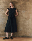 A woman with wavy hair and a relaxed expression stands against a textured beige wall wearing The Tulsa Dress by The Great Inc., styled with black oxford shoes and a vintage bandana headband.