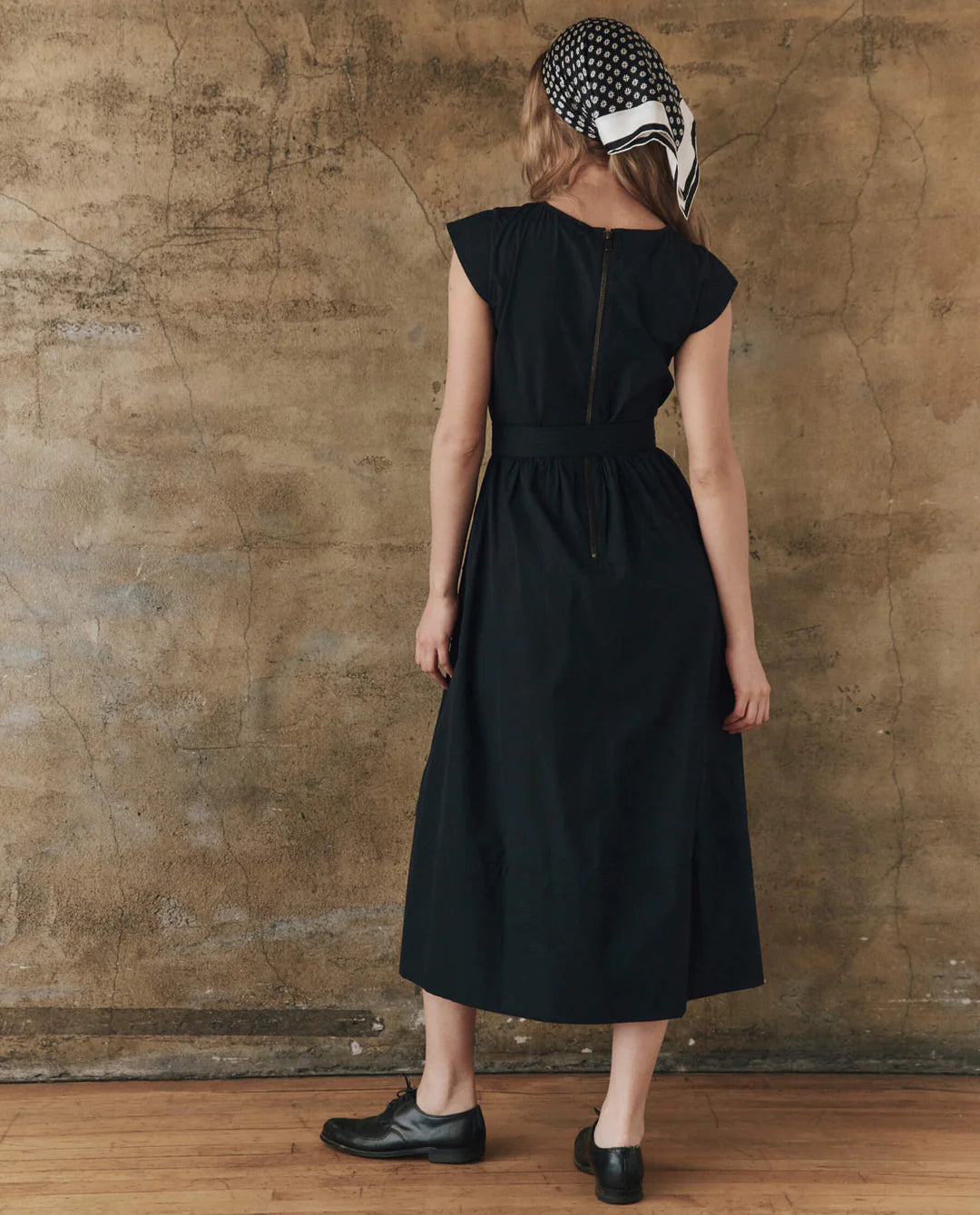 A woman stands with her back to the camera in The Great Inc.'s Tulsa Dress, styled with black shoes and a vintage bandana headscarf, set against a tan textured wall and wooden floor.
