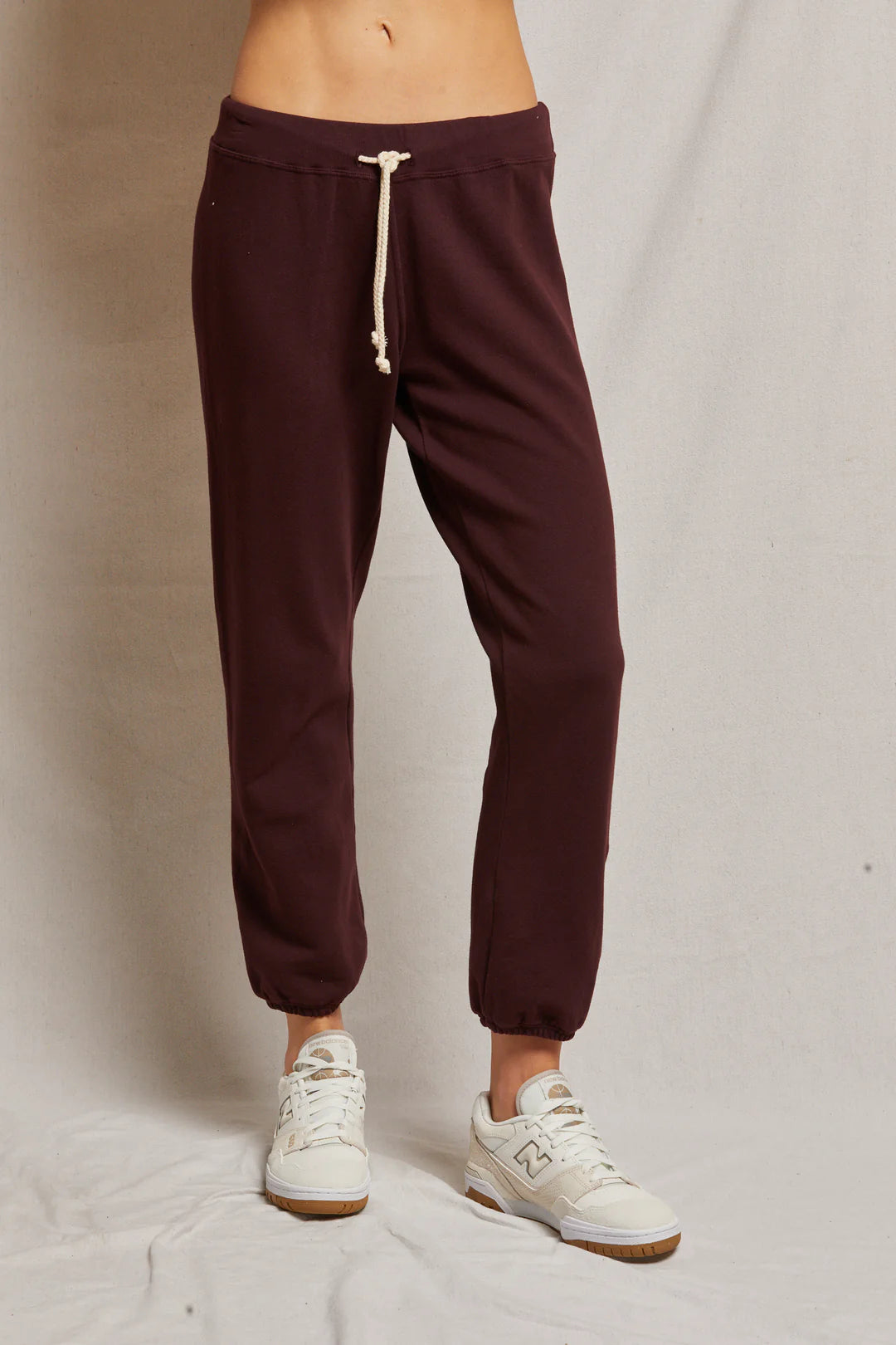 A person wearing the Toni French Terry Jogger by Perfectwhitetee in maroon and white sneakers with subtle brown accents stands against a plain beige background. The joggers feature an adjustable drawstring, and only the lower half of the body is visible.