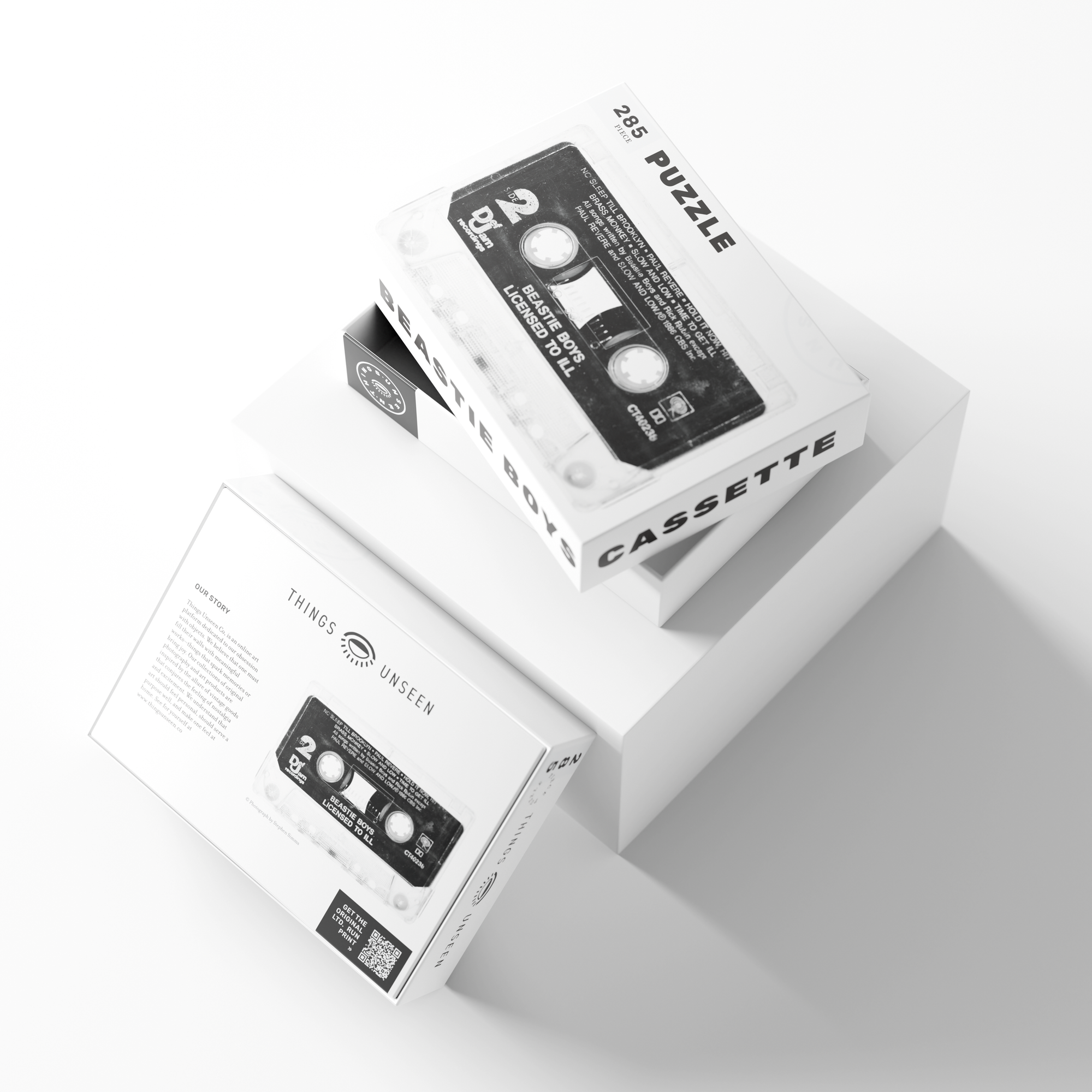 A jigsaw puzzle box with a black and white design showcases an image of a cassette tape that pays tribute to the Beastie Boys&#39; debut LP. The box, labeled &quot;PUZZLE - BEASTIE BOYS - LICENSED TO ILL,&quot; sits on white geometric shapes and features the brand name &quot;Things Unseen&quot; on its side.