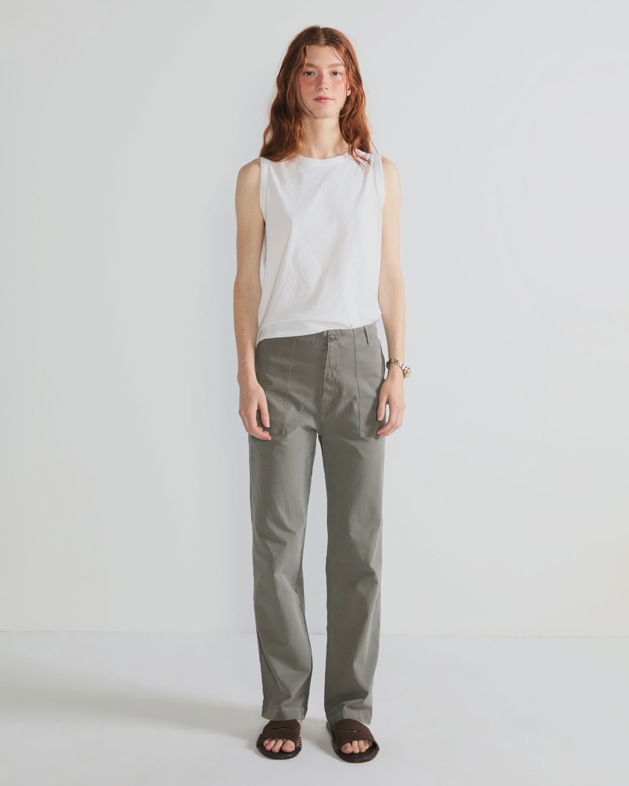 A person with long reddish hair wears Kule’s The Tank, paired with gray straight-leg pants and black sandals, standing against a plain white background.