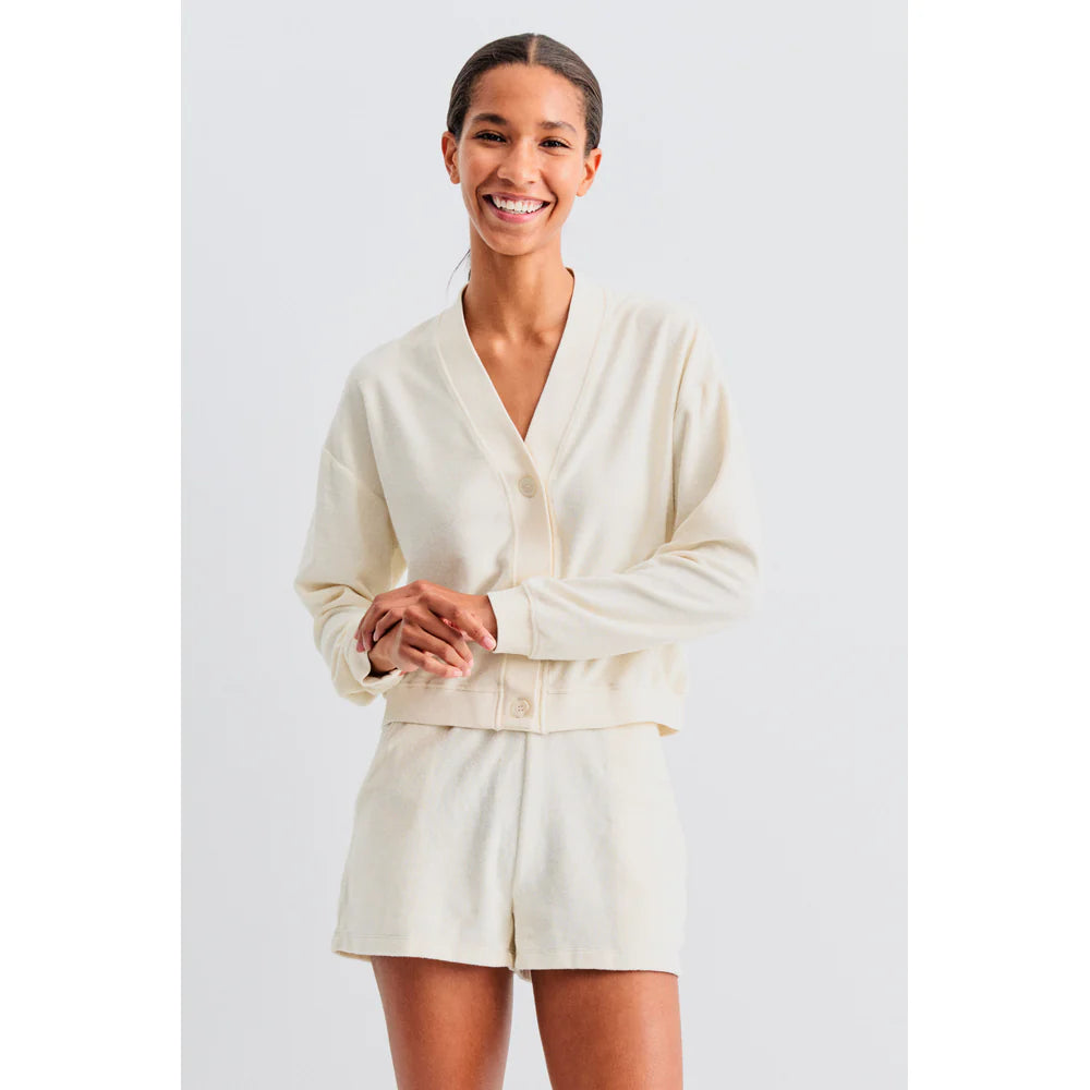 A person smiles in Kule's The Terry Bay, a relaxed fit, cream-colored cardigan with matching shorts, exuding casual style. The plain white background enhances the clothing's ease and coziness.