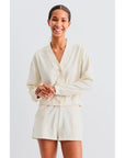 A person smiles in Kule's The Terry Bay, a relaxed fit, cream-colored cardigan with matching shorts, exuding casual style. The plain white background enhances the clothing's ease and coziness.