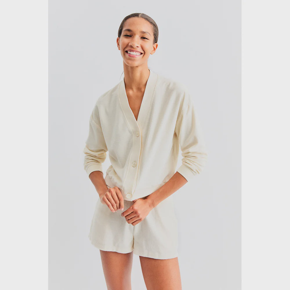 A person is smiling and wearing The Terry Bay set by Kule, which includes a light cream long-sleeve buttoned top resembling a cropped cardigan paired with matching shorts. The background is plain white.