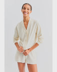 A person is smiling and wearing The Terry Bay set by Kule, which includes a light cream long-sleeve buttoned top resembling a cropped cardigan paired with matching shorts. The background is plain white.