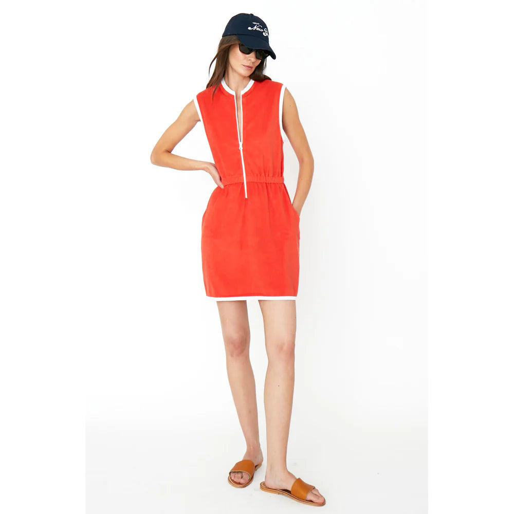 A person is wearing The Terry Dress by Kule, a chic sleeveless red dress with a front zipper, white trim, and pockets. It's paired with a black cap, sunglasses, and brown slide sandals against a plain white background.