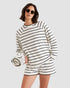 A woman wearing the Kule Terry Franny sweatshirt with matching striped shorts and sunglasses holds a rolled-up striped towel against a plain light gray background.