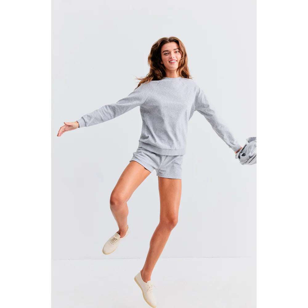 A person jumps joyfully in a light gray sweatshirt and Kule's Terry Venus shorts with an elastic waist, holding a gray jacket. They wear light-colored shoes, all against a plain white background.