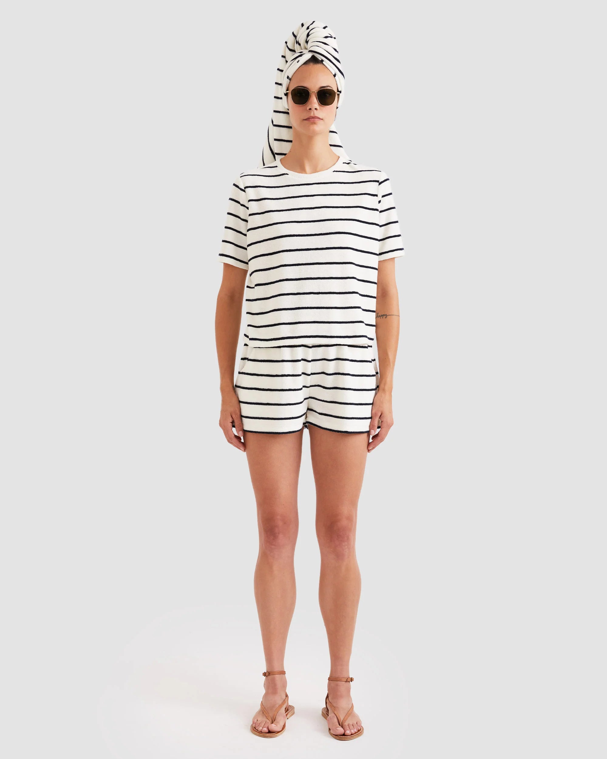 A person stands facing forward against a plain background, wearing sunglasses, a white striped T-shirt, and Kule's The Terry Venus short—a cotton-blend style with an elastic waist. A towel is wrapped around their head; they are barefoot and neutral-faced.