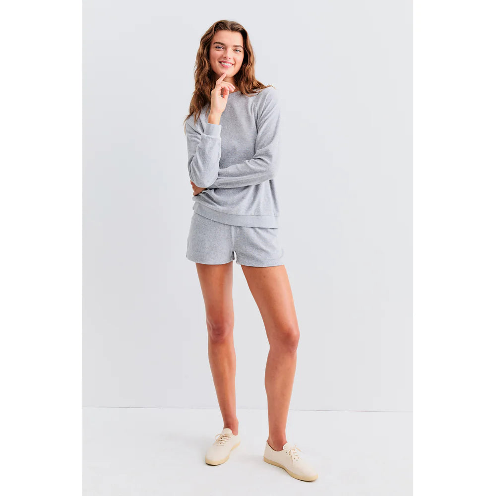 A long-haired person smiles with one hand on their chin, wearing Kule's The Terry Franny sweatshirt in light grey and matching shorts, paired with light-colored shoes against a plain white background.