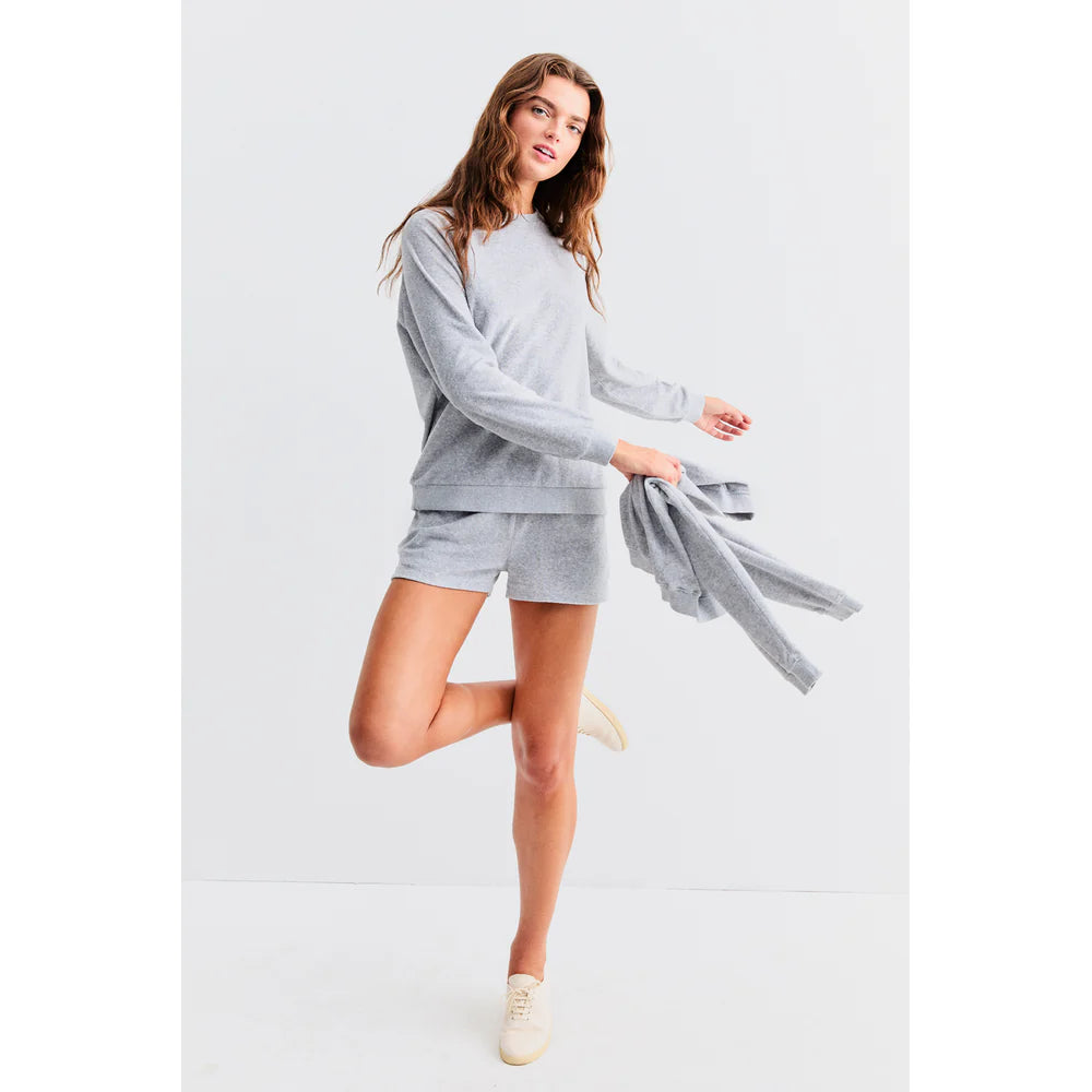 A person stands joyfully on one leg against a light background, wearing Kule's The Terry Franny sweatshirt and shorts. They hold a matching top in one hand, smiling in light sneakers. The outfit's casual charm is enhanced by the raglan sleeves of the gray cotton-blend sweater.