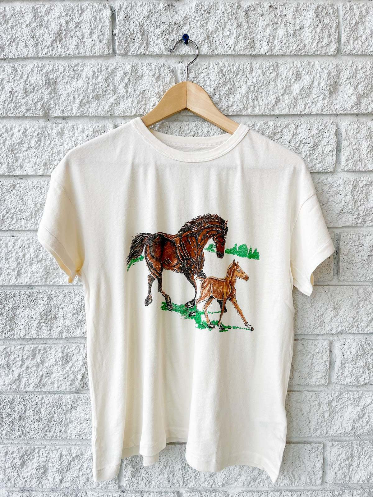 The Boxy Crew W/ Running Horses by The Great Inc. is a cream-colored, boxy-fit t-shirt shown on a wooden hanger against a white brick wall, featuring detailed graphics of a brown horse and foal running on green grass.
