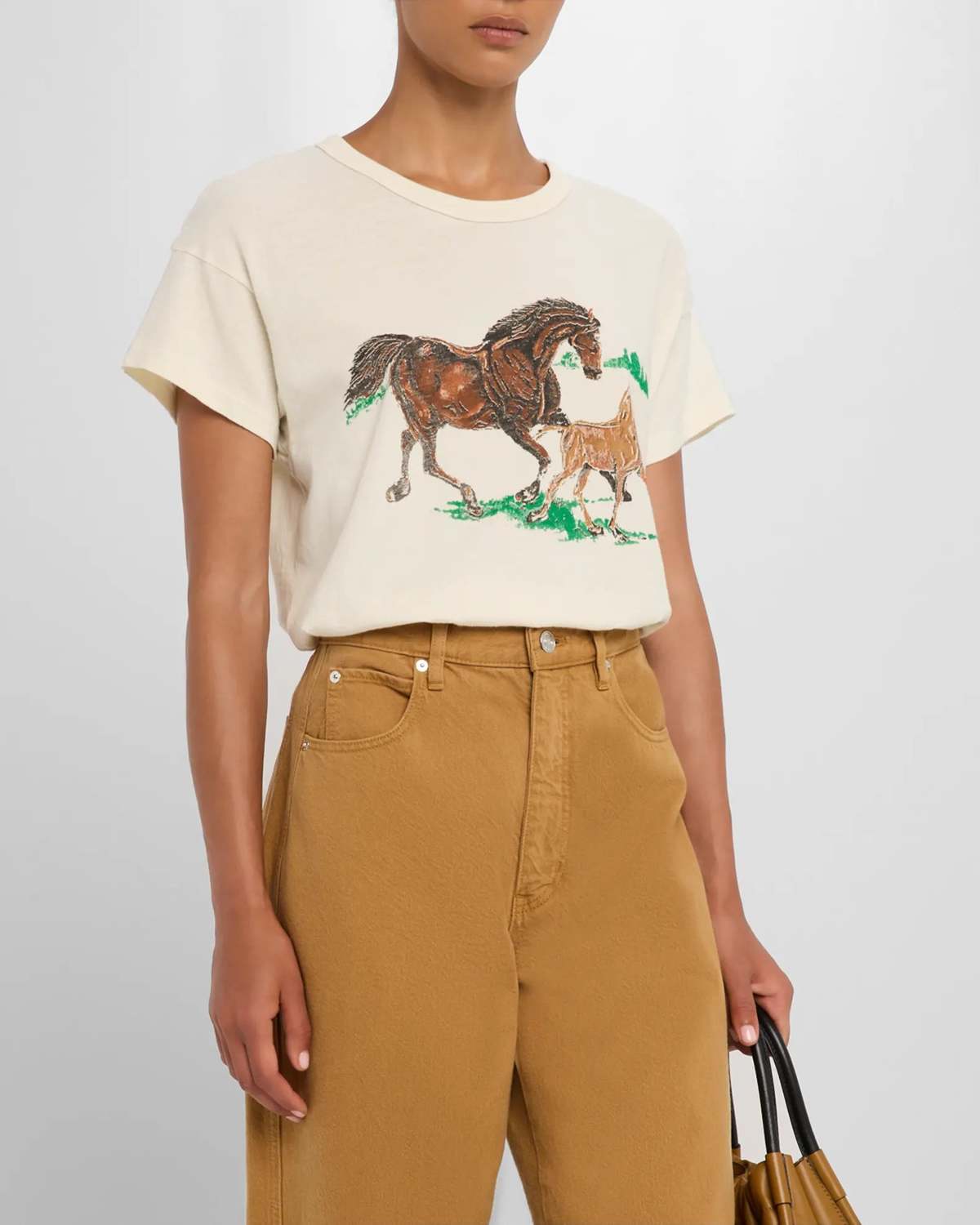 Wearing The Great Inc.'s Boxy Crew W/ Running Horses, paired with tan high-waisted pants and holding a brown handbag at their side.