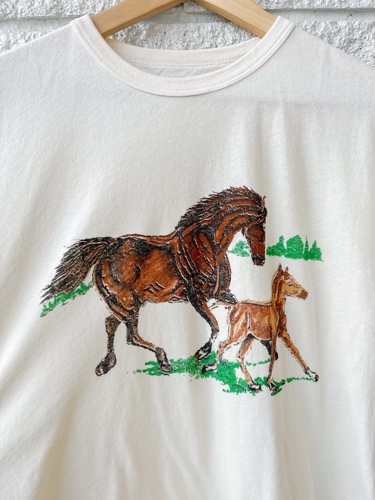 The Boxy Crew W/ Running Horses by The Great Inc. is a white crew neck tee featuring brown adult and foal horses trotting on green grass, with subtle horse details and faint trees in the background.