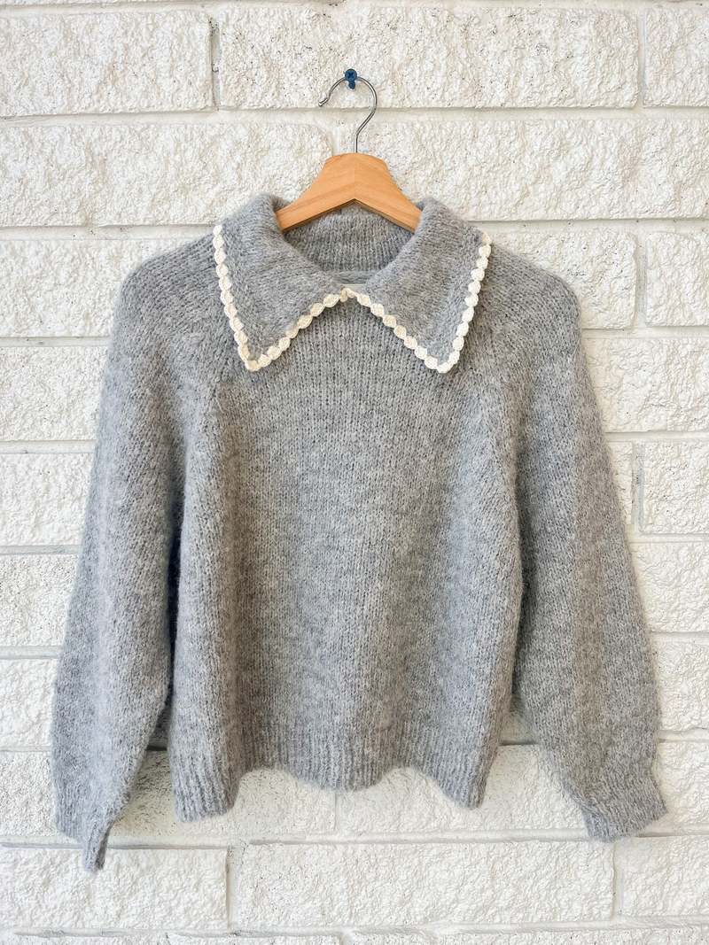 The Great Inc.'s Crochet Collar Pullover is a gray knit, long-sleeve sweater with a scalloped white collar, displayed on a wooden hanger against a white brick wall.