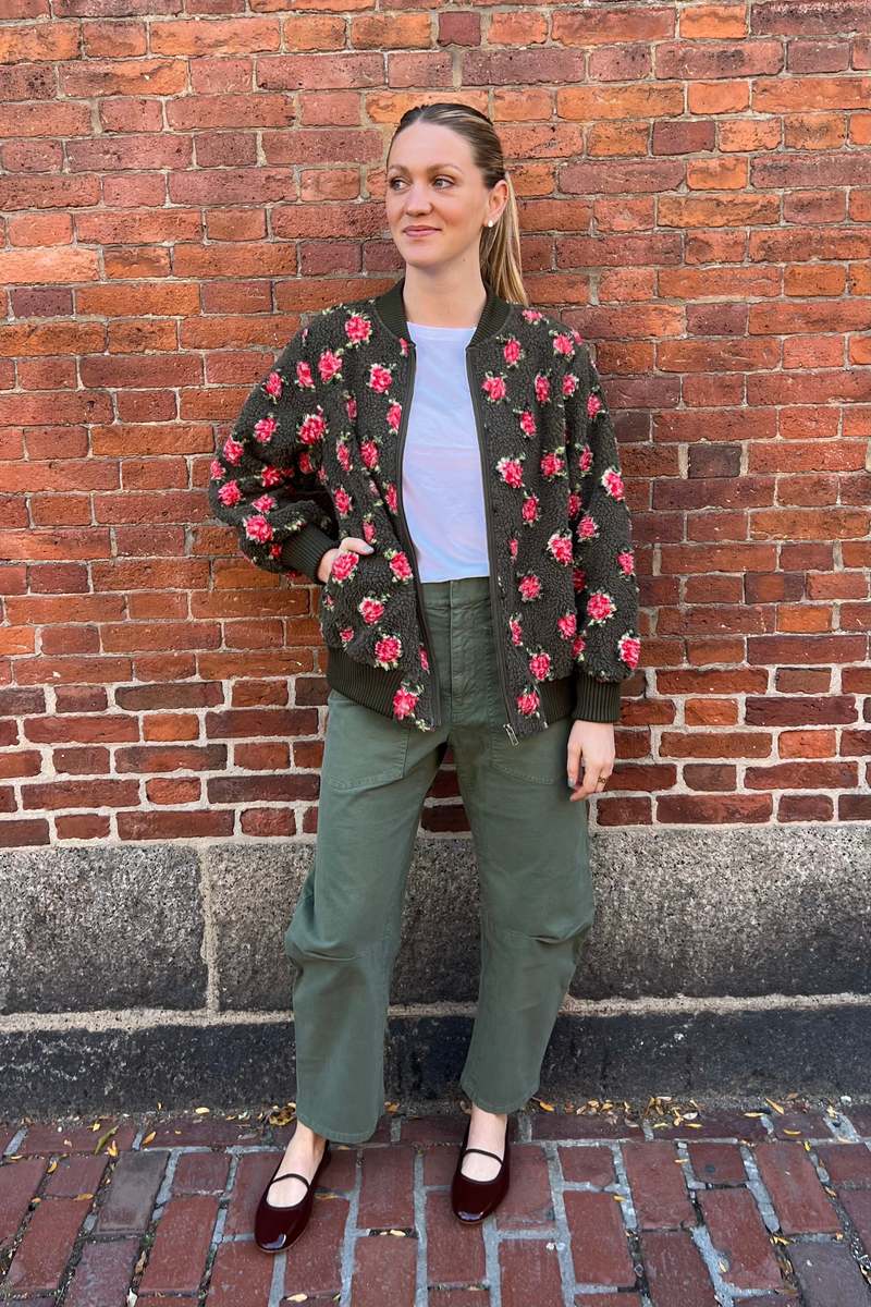 A person stands before a brick wall, wearing The Great Inc.'s "The Zip Front Plush Bomber" in army green with a rose pattern, styled with a white tee, green pants, burgundy flats; hair tied back and one hand on their hip.