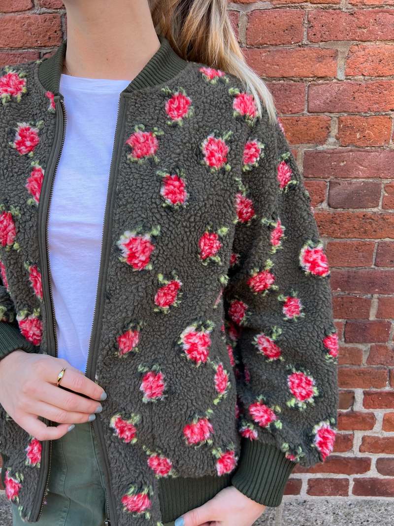 A person stands against a brick wall wearing The Great Inc.'s The Zip Front Plush Bomber in army green with a red and white rose pattern, paired with a white T-shirt and green pants, hand near the zipper.