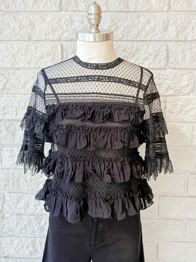 The Winter Mesh Flounce Top by The Great Inc. is a black sheer lace blouse with a sheer upper bodice, horizontal ruffled layers, and intricate woven lace details, displayed on a mannequin against a white brick wall.