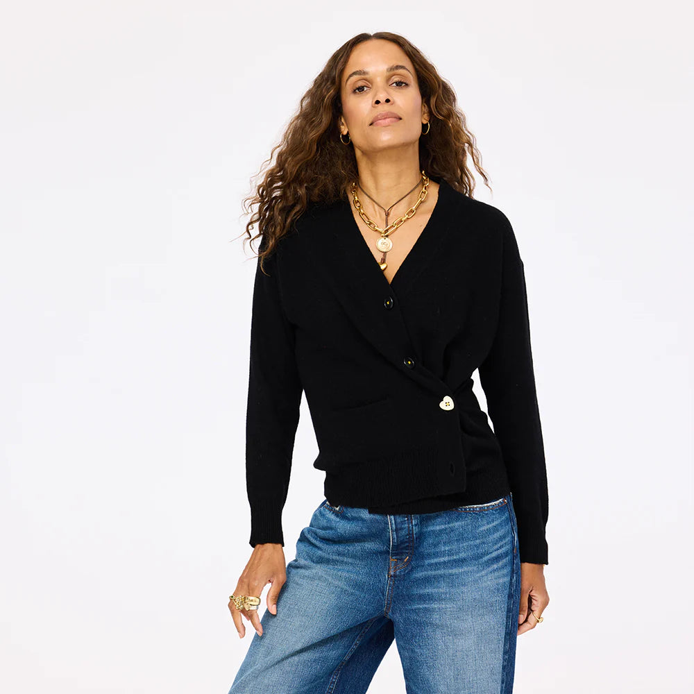 A woman with long curly hair wears the Jackie Cardigan by Kerri Rosenthal—a black V-neck cardigan with buttons—paired with blue jeans and layered gold necklaces, standing confidently against a plain white background.
