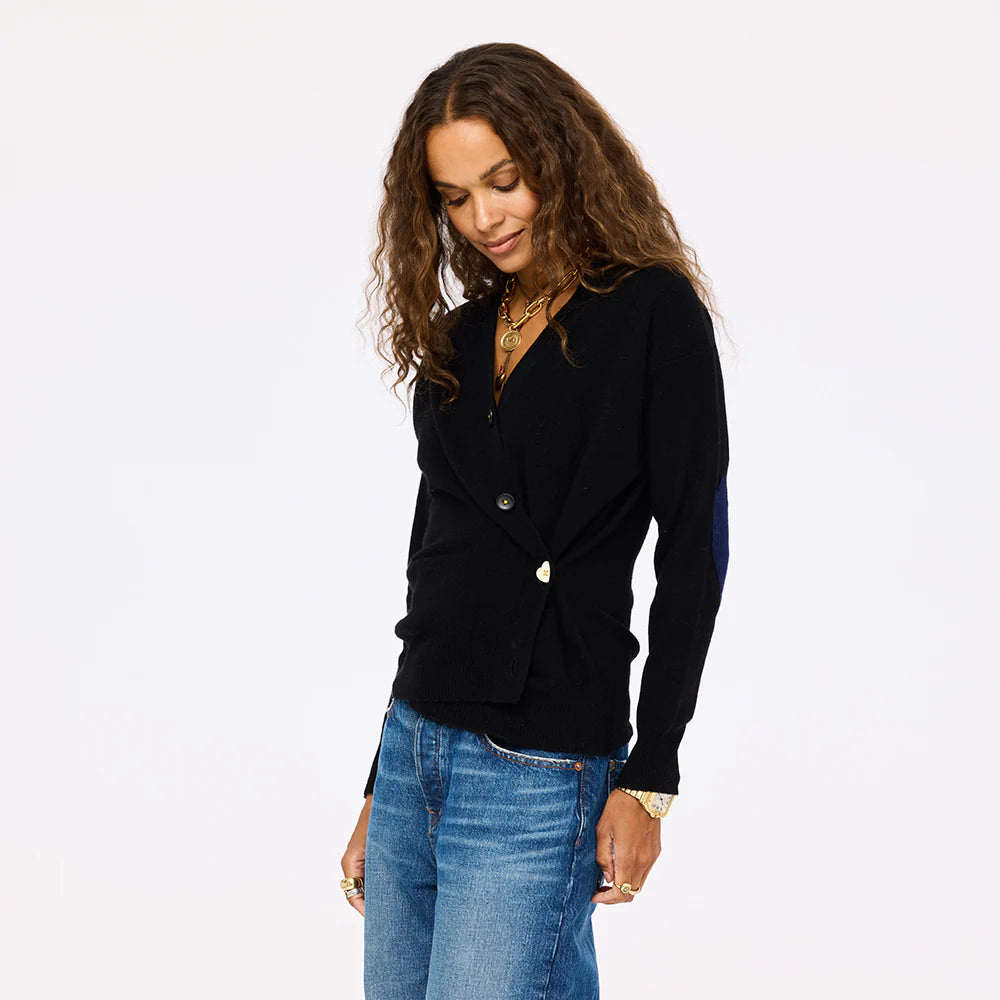 A woman with long curly hair, wearing the Kerri Rosenthal Jackie Cardigan in black and blue jeans, stands against a plain light background, looking down with a relaxed expression.
