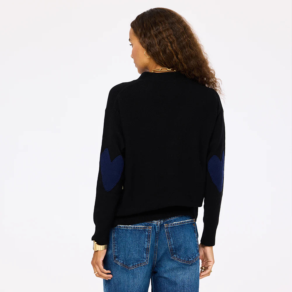 A woman with curly brown hair, wearing the Kerri Rosenthal Jackie Cardigan—black with blue heart-shaped elbow patches—and blue jeans, stands facing away against a plain light background.