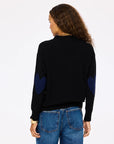 A woman with curly brown hair, wearing the Kerri Rosenthal Jackie Cardigan—black with blue heart-shaped elbow patches—and blue jeans, stands facing away against a plain light background.